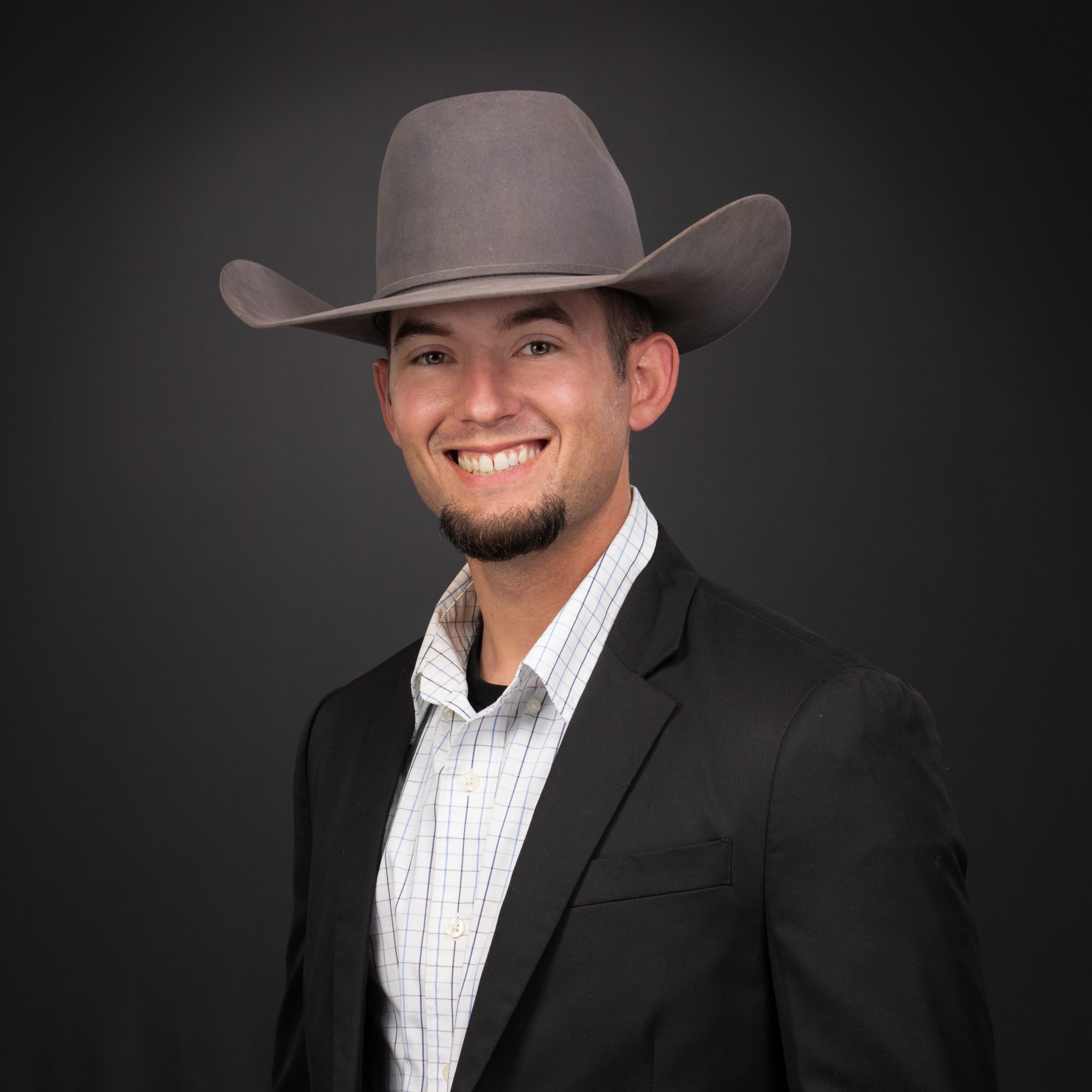 Business headshot of a smiling man in a gray cowboy hat, black suit jacket and checked shirt on a dark gray backdrop