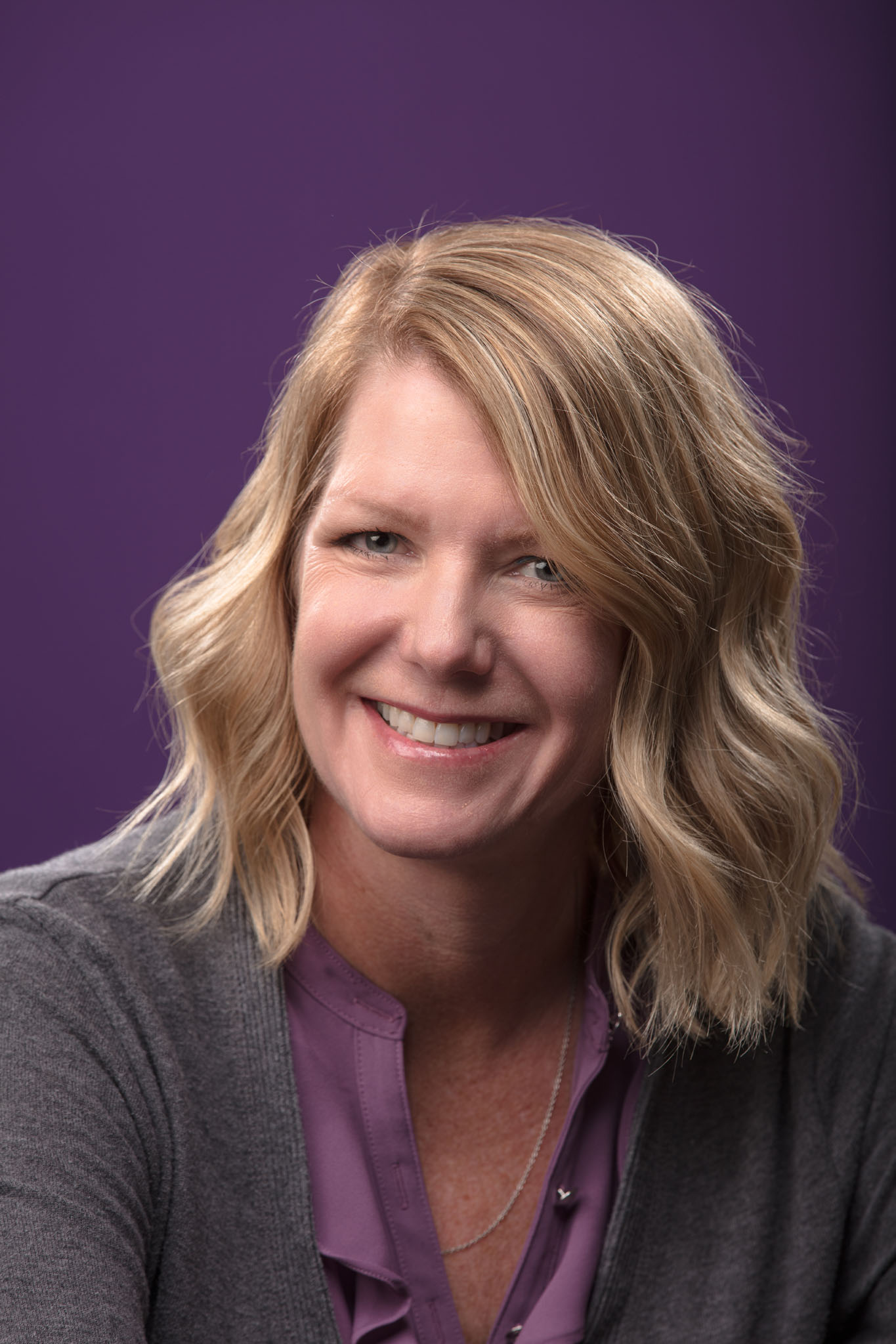 Professional headshot of a smiling woman with wavy blonde hair in a gray cardigan and purple top against a purple backdrop