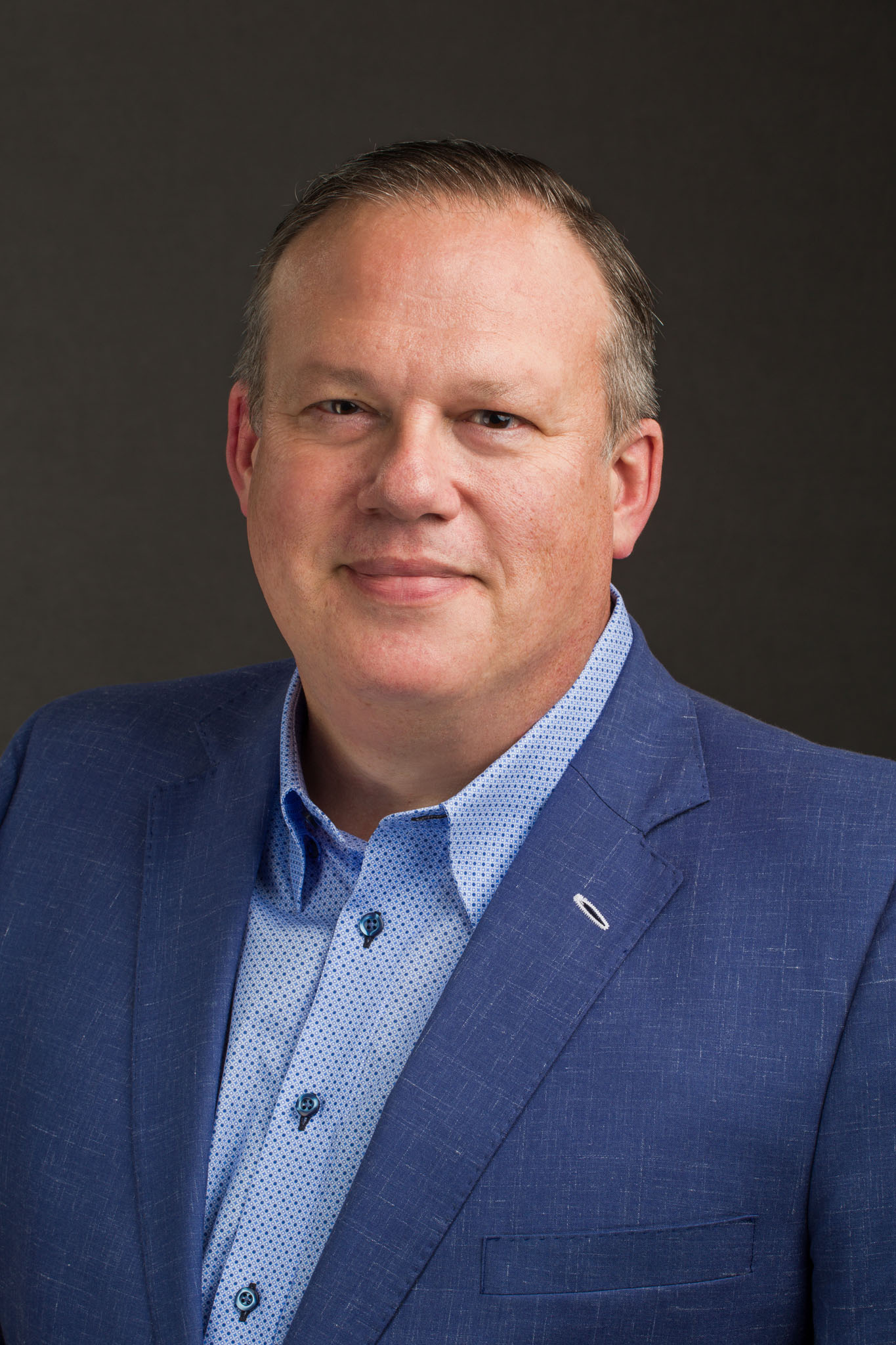 Executive headshot of a smiling man in a blue sport coat and patterned blue shirt on a dark gray backdrop