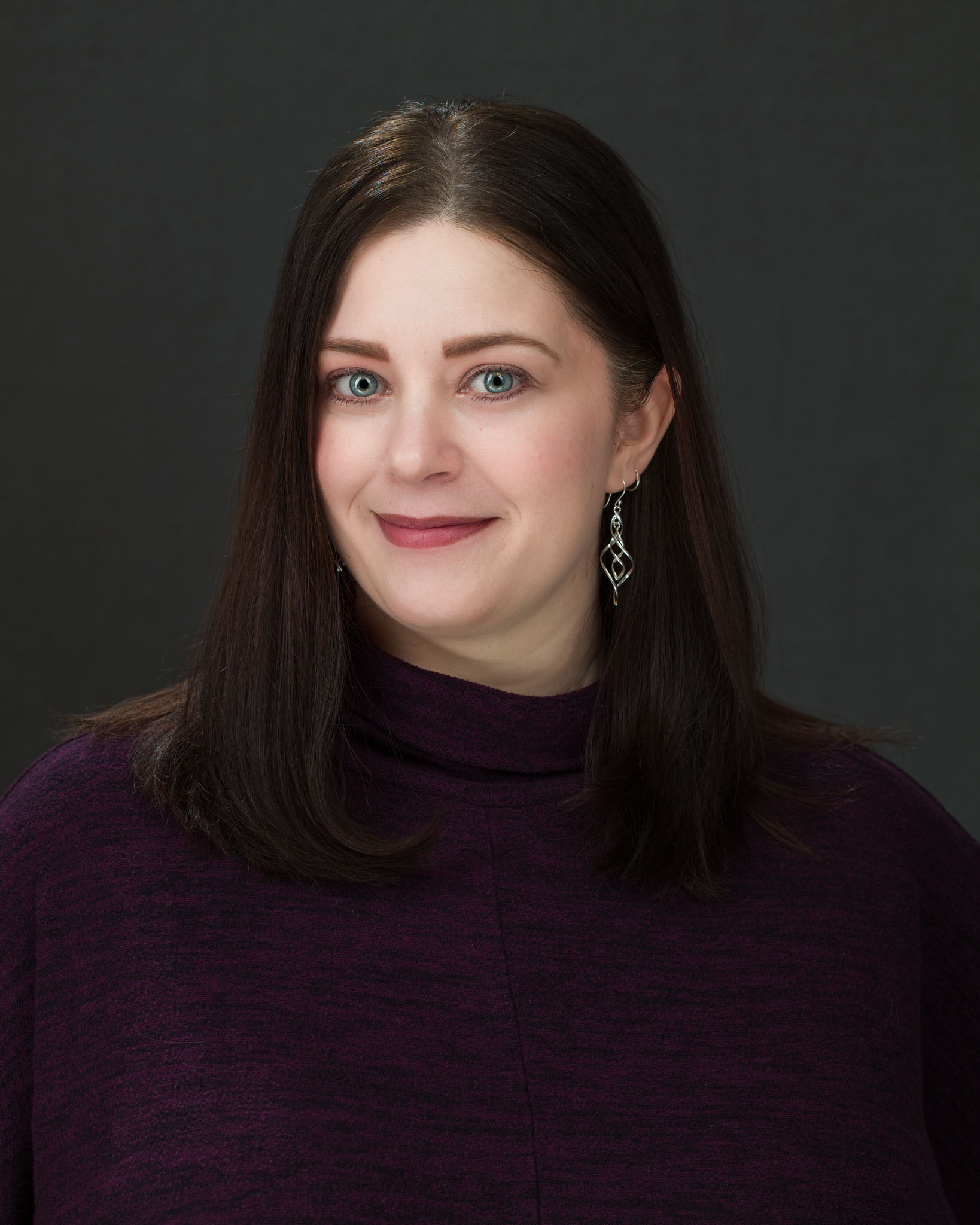Professional headshot of a woman with dark shoulder-length hair in a plum sweater on a dark gray backdrop