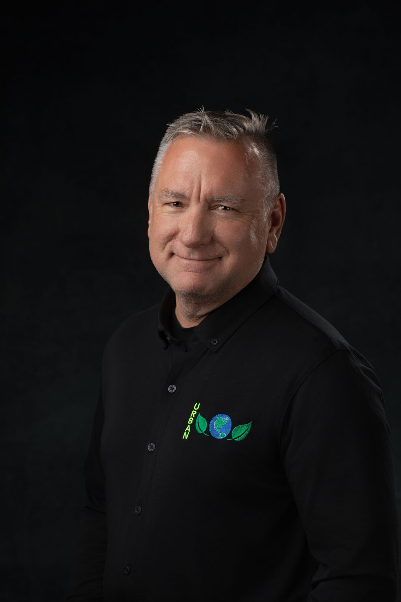 Business headshot of a smiling man with gray hair in a black logo shirt on a black backdrop