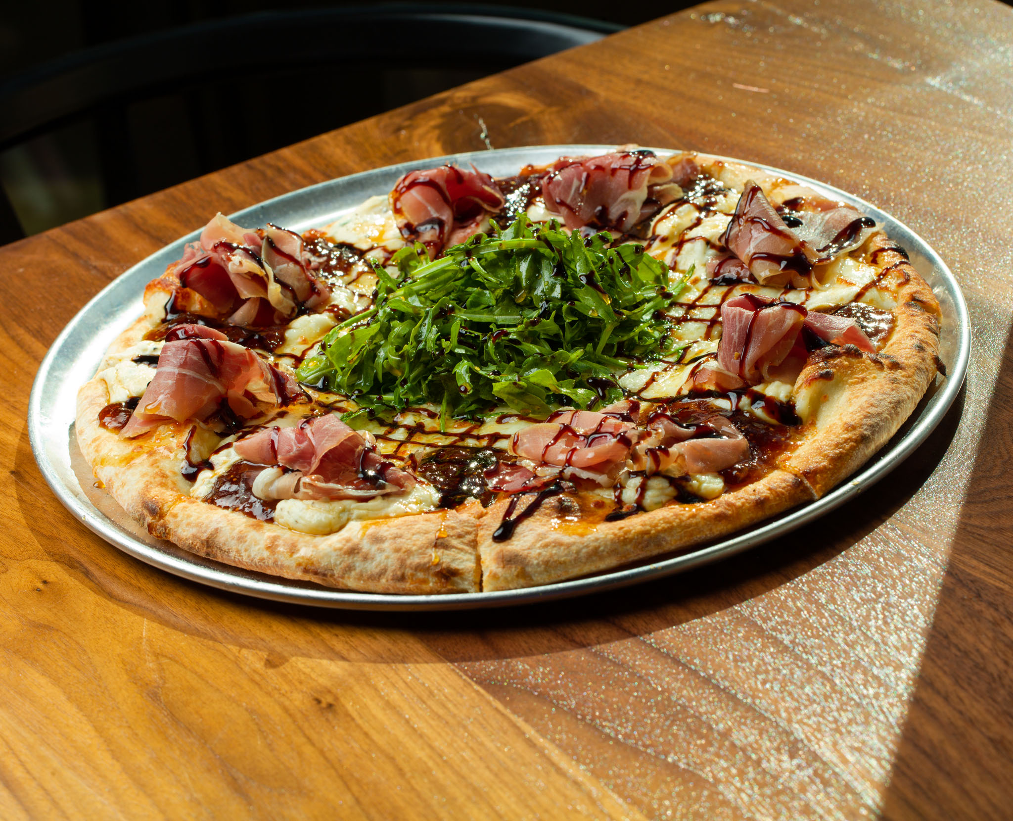 Pizza topped with prosciutto, a mound of arugula and a balsamic drizzle on a metal tray on a wooden table in afternoon sun