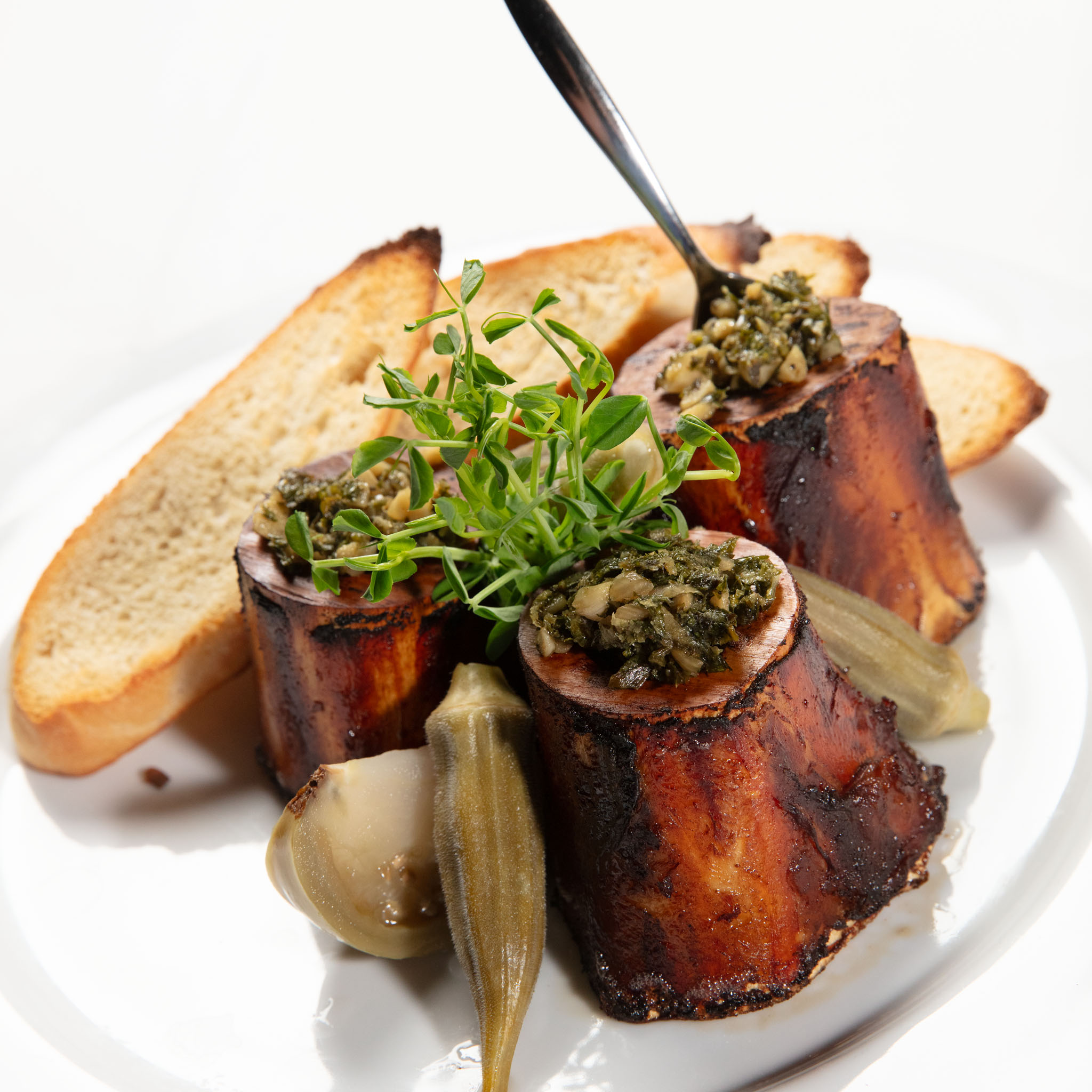 Roasted marrow bones topped with a green herb relish, served with toasted bread, pickled okra and a sprig of herbs on a white plate