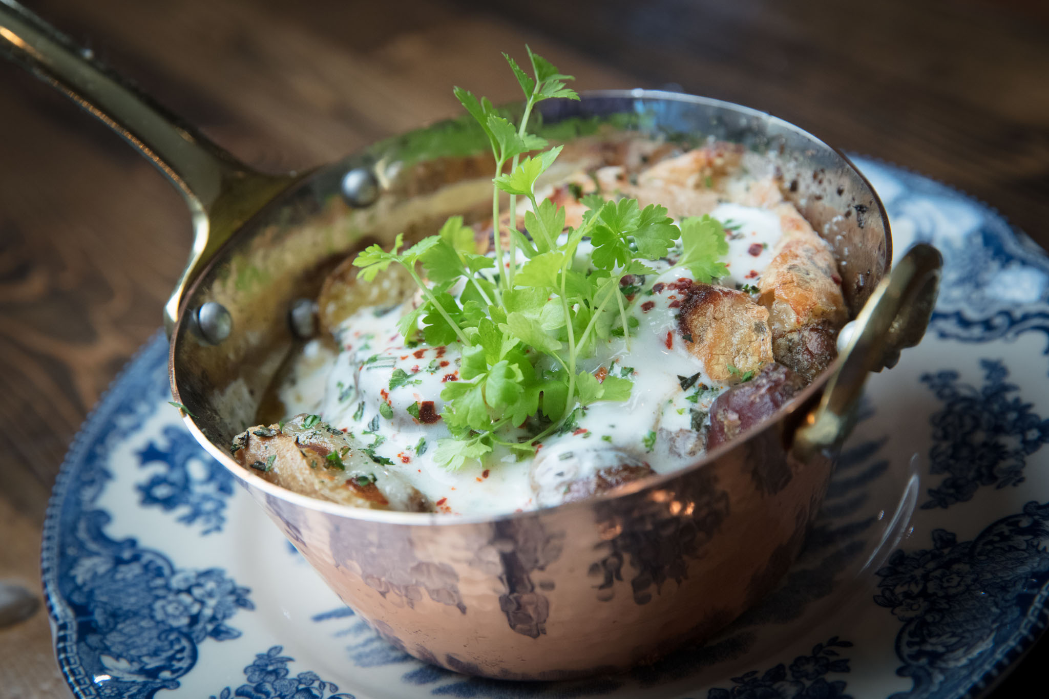 Small copper pan of roasted potatoes in white yogurt sauce with chili flakes and fresh herbs, set on a blue-and-white patterned plate
