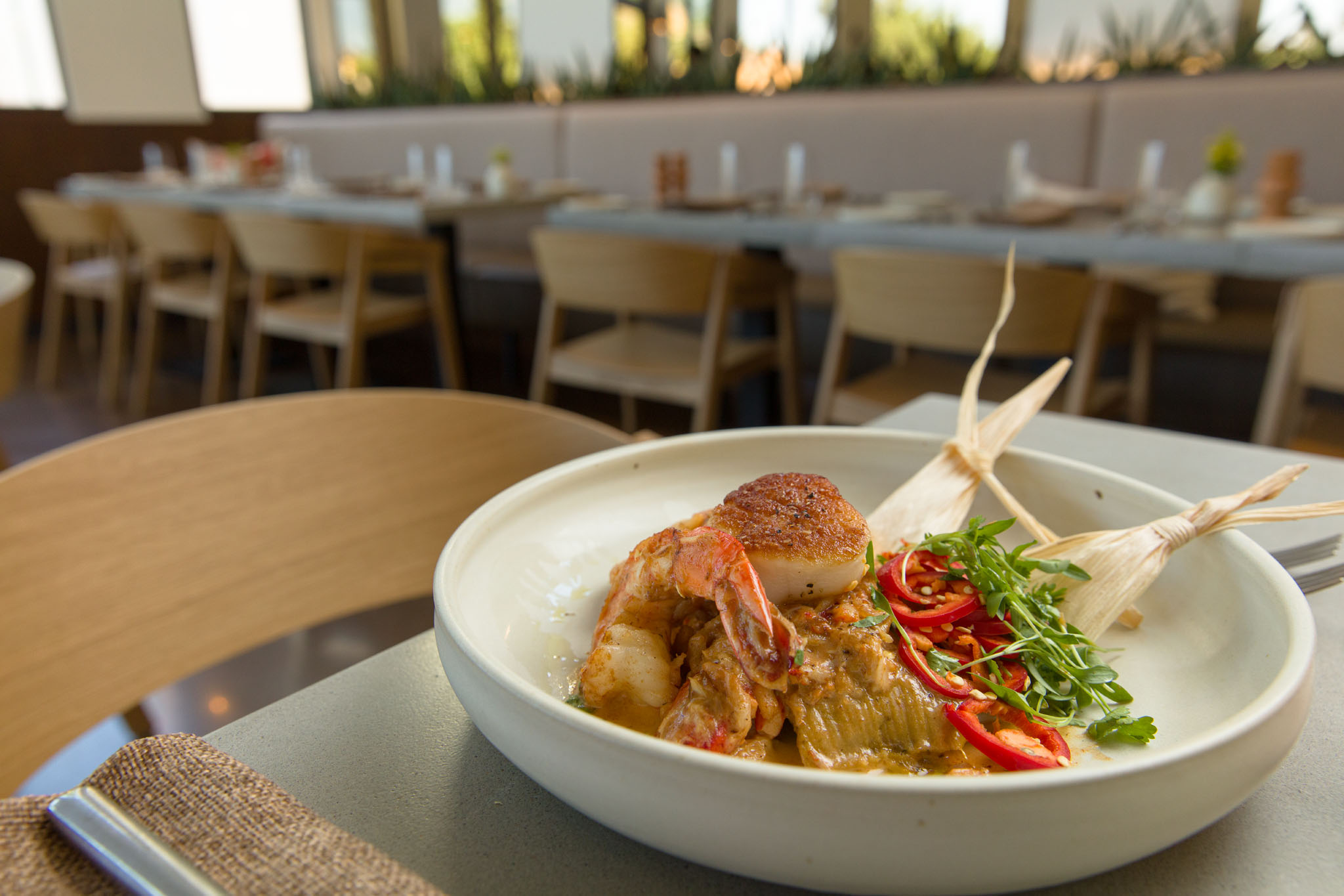 Bowl of shrimp and seared scallop in a creamy sauce with sliced red chilies, microgreens and corn husks, with a restaurant dining room behind