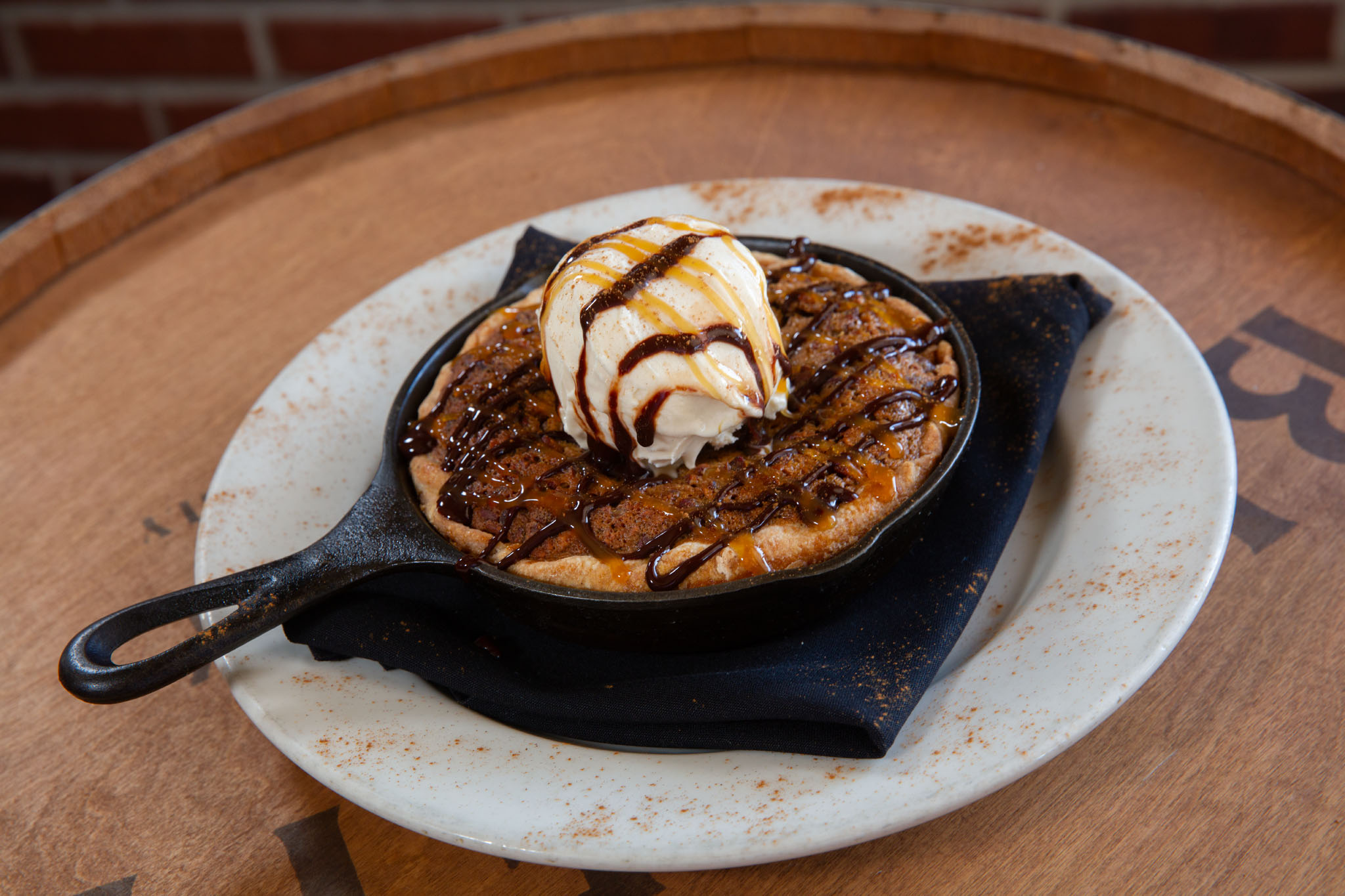 Warm skillet dessert topped with a scoop of ice cream and caramel and chocolate drizzle, served on a plate over a dark napkin on a barrel top