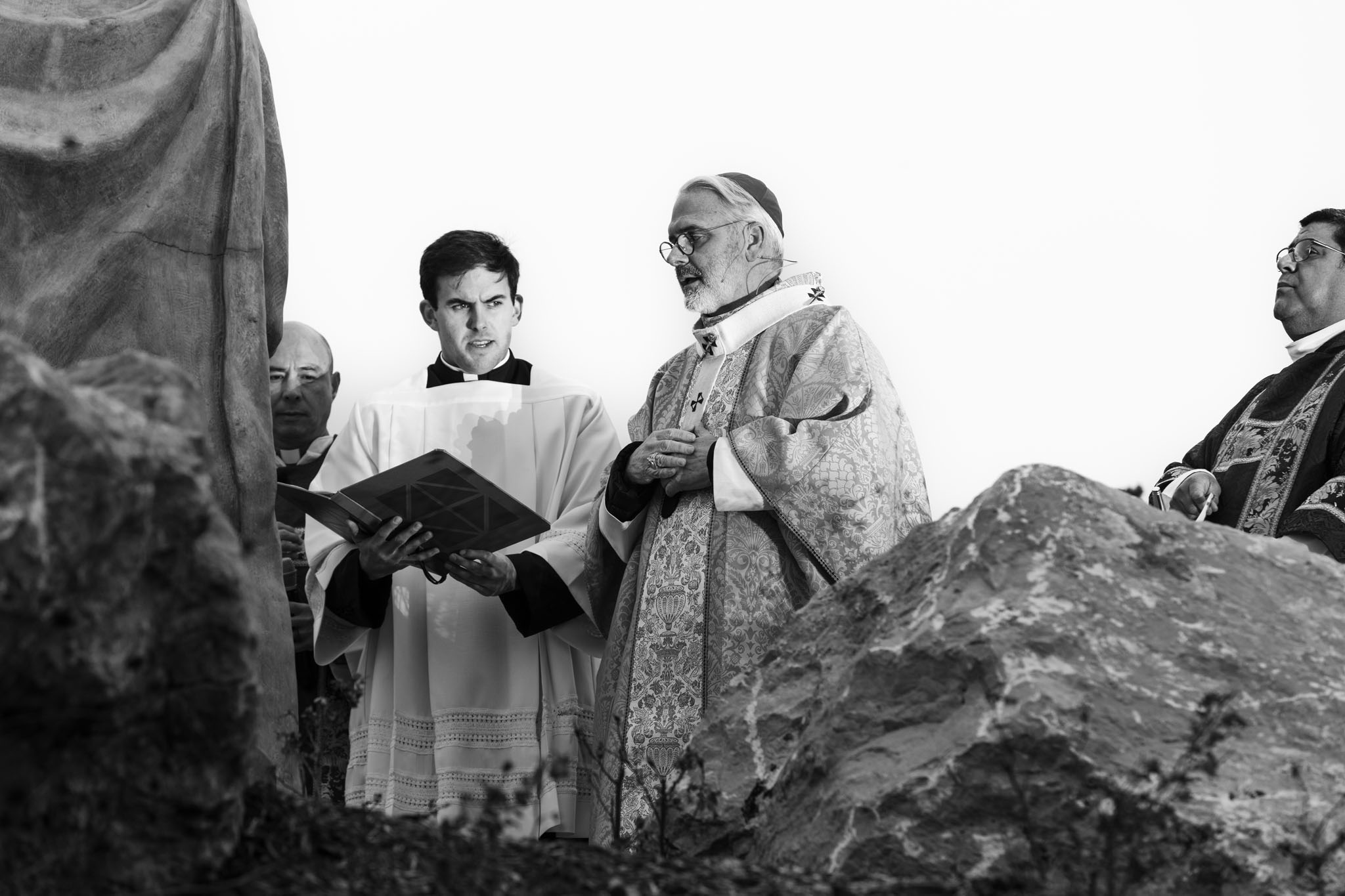 Bishop reading a prayer beside altar servers among the rocks
