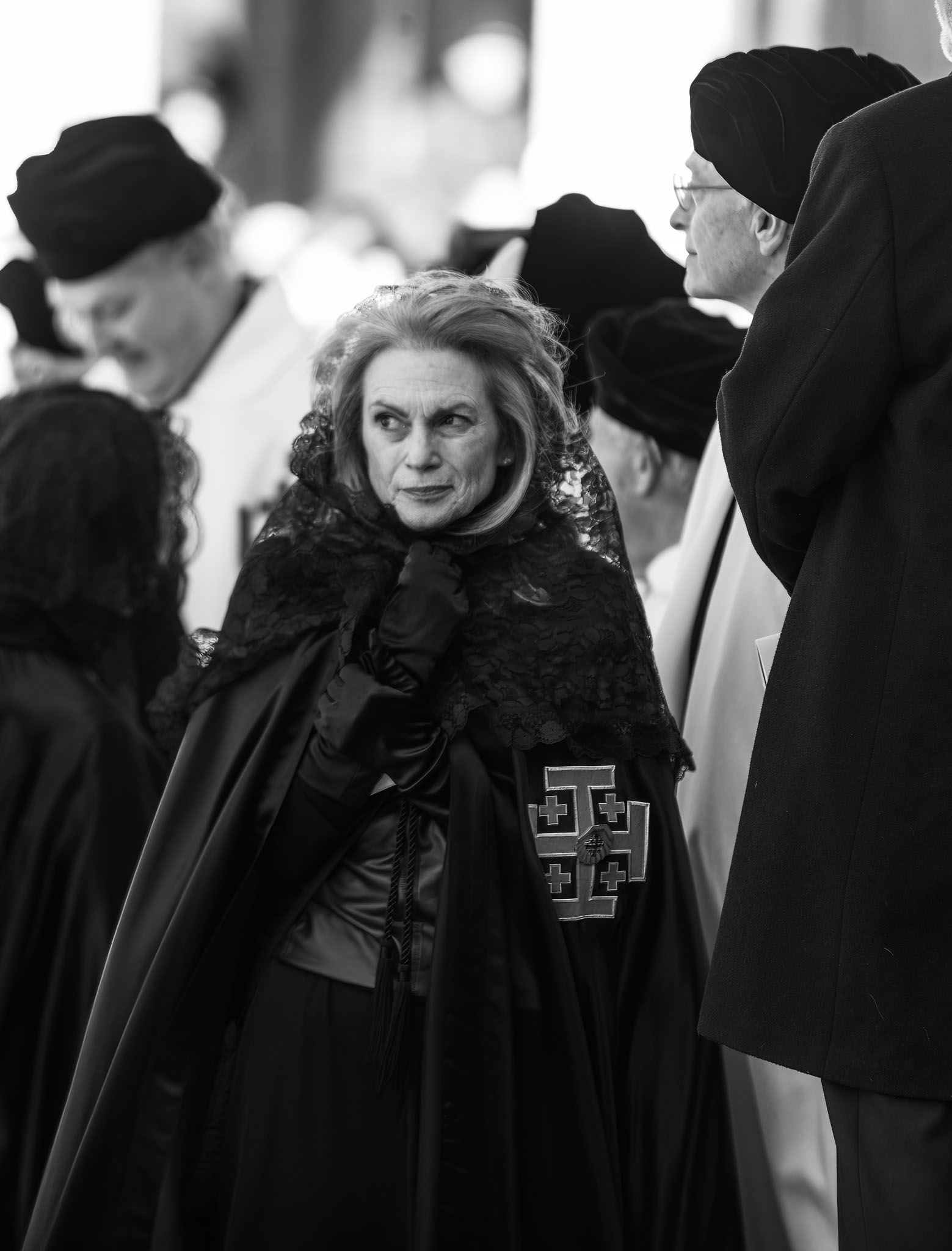 Woman in a dark cloak with a cross emblem among the crowd