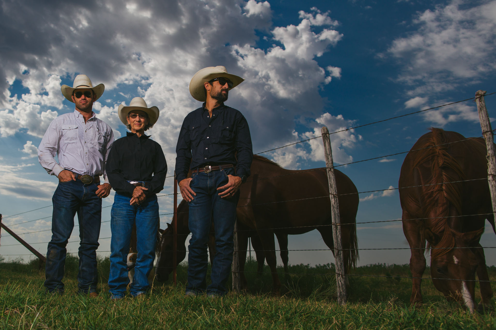 Three cowboys standing at a fence line with horses under a cloudy sky