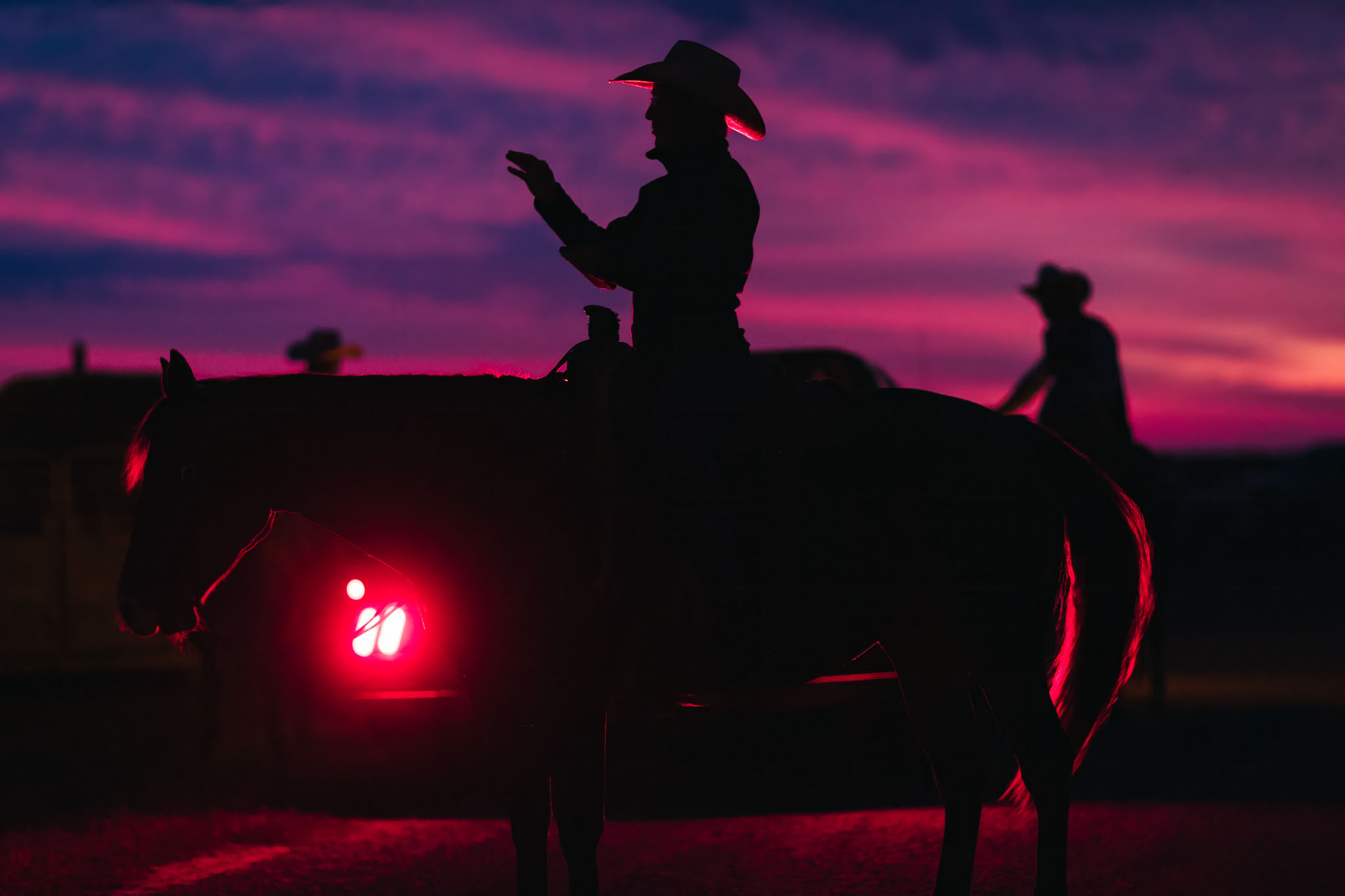 Cowboy on horseback silhouetted against a pink and purple dawn sky, a truck’s red light glowing below
