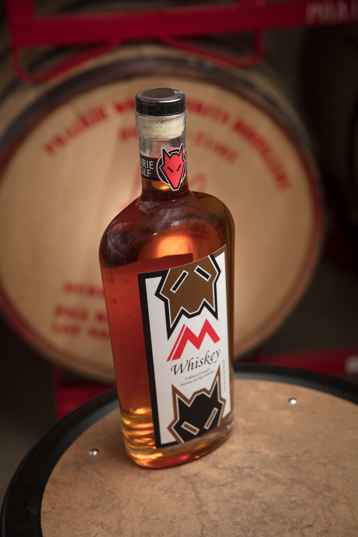 Bottle of whiskey with a wolf-themed label standing on the head of a whiskey barrel, with another barrel behind it